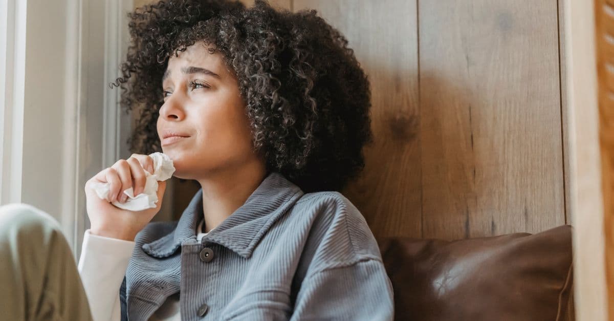 Unhappy young African American female with dark curly hair in casual clothes crying and looking away while sitting near window at home