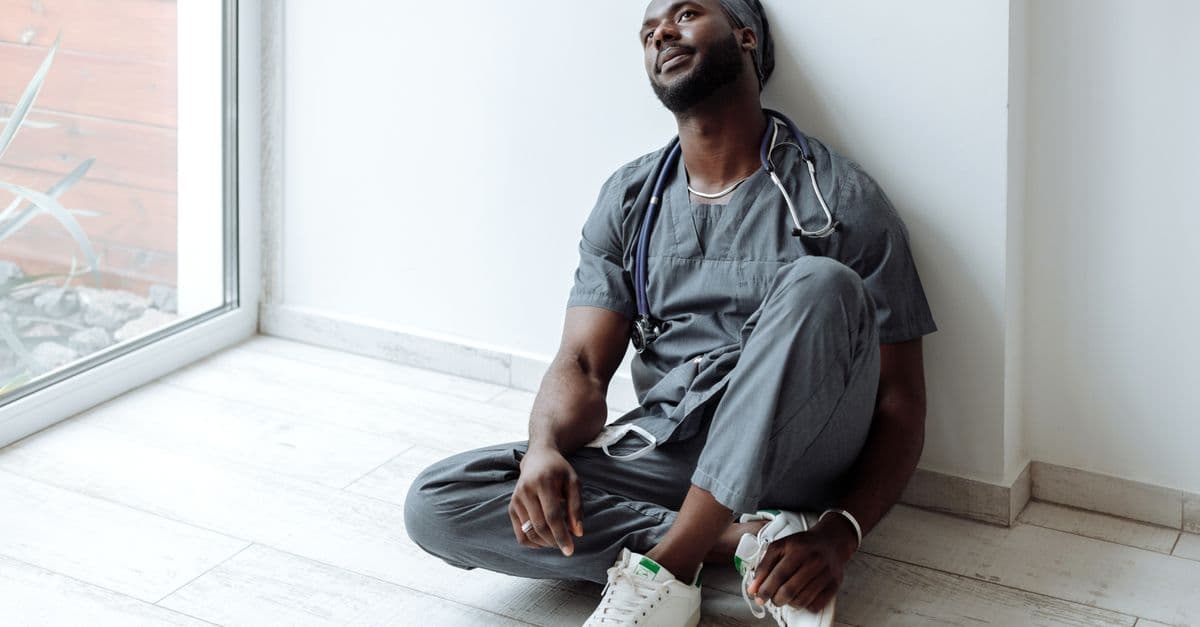 A tired healthcare professional in gray scrubs resting against a wall, reflecting exhaustion.
