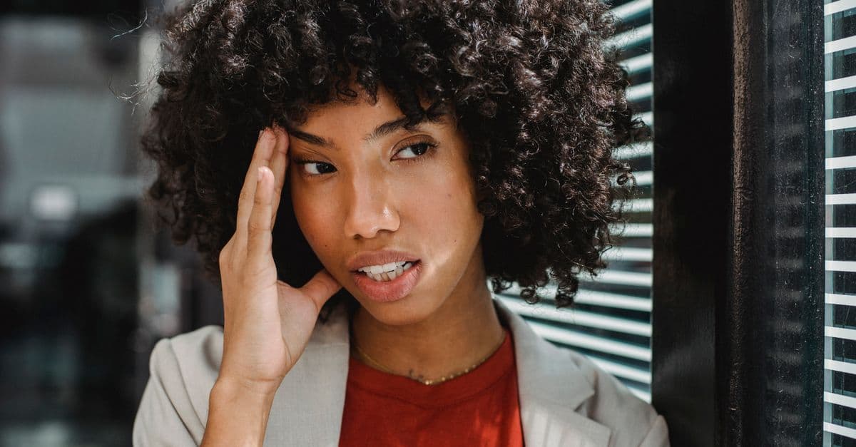A businesswoman with curly hair expresses stress by her office window, touching her forehead.