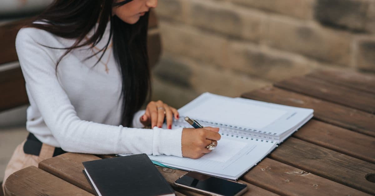Crop ethnic female manager taking notes in notepad while working at wooden table with smartphone and diary