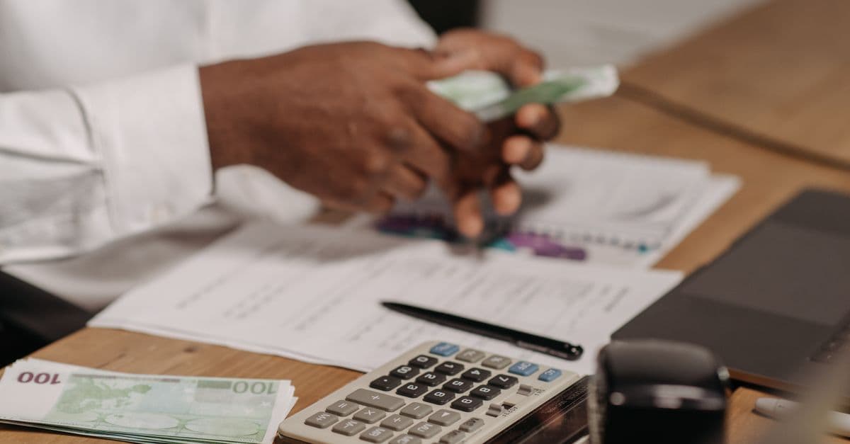 Hands counting euro bills on a wooden desk with calculator, financial documents, and laptop nearby.