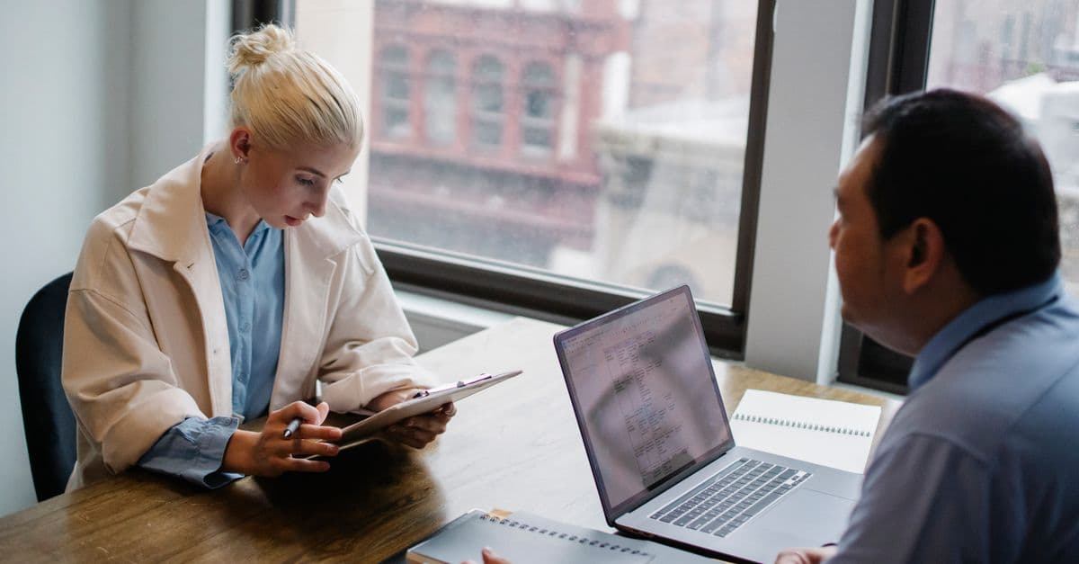 From above of young female worker wearing modern coat analyzing details of project with colleague while sitting together at wooden desk with laptop and documents
