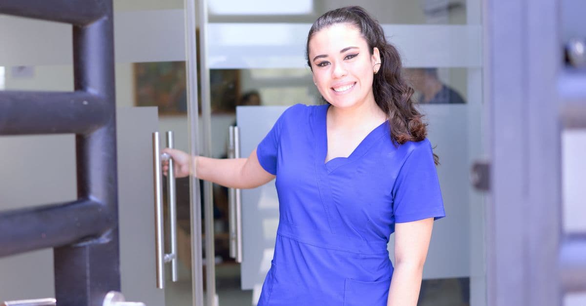 Smiling female nurse in blue scrubs opens a glass door in a healthcare setting.