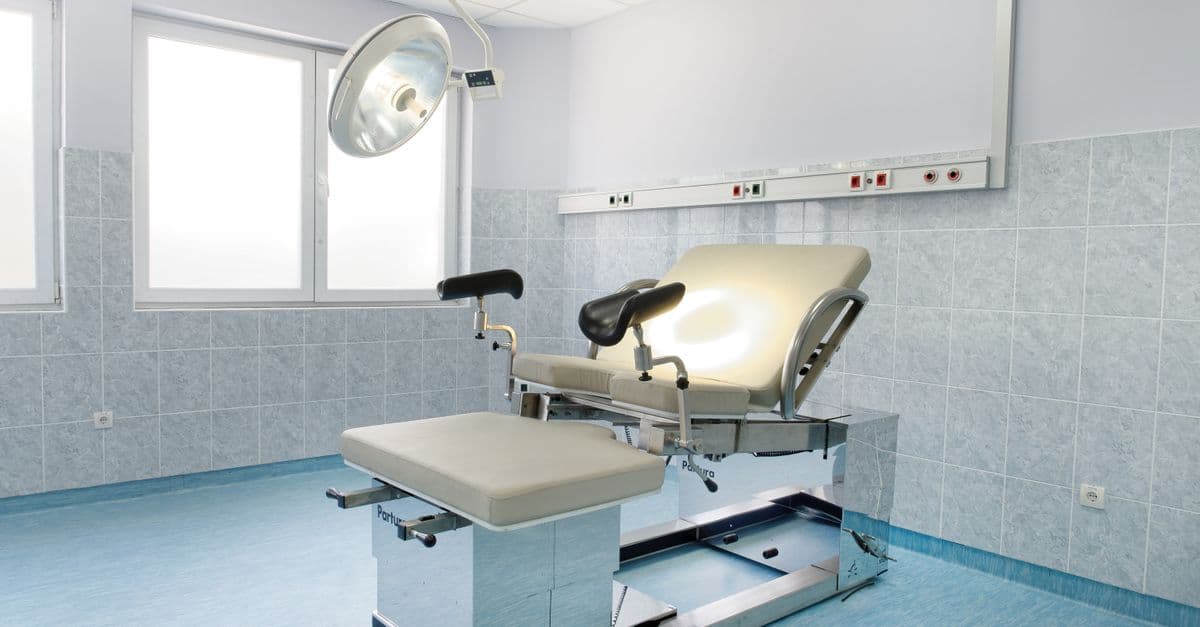 Bright gynecological examination room with adjustable chair and overhead light in a medical facility.